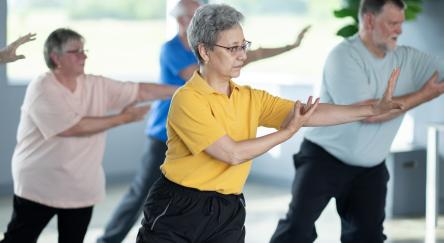 A small group of seniors are seen practicing Tai Chi in an indoor studio. They are each dressed comfortably and are focused on their movements and proper form as they practice the Martial Art.