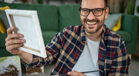 A smiling man in a casual plaid shirt enjoys the moment while painting on a canvas.