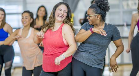 A multi-ethnic group of adult women are dancing in a fitness studio. They are wearing athletic clothes.