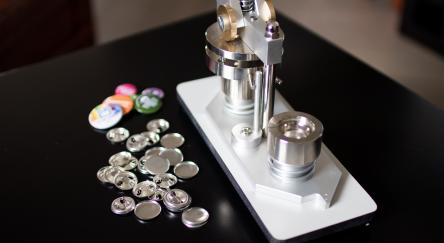 A button making machine sits on a table with pins and covers sitting nearby.