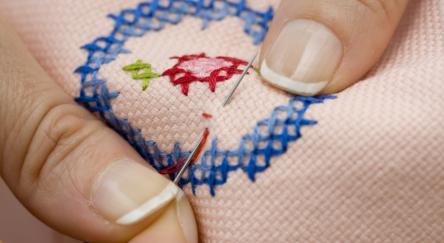 Close up of hands doing a cross stitch pattern on salmon coloured cloth.