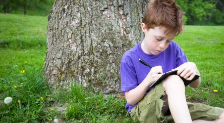 Little boy sitting in grass outside, writing in a notebook or journal.
