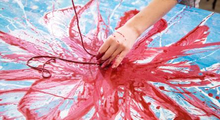 Close up of female painter bare hand creating a painting on canvas lying on the table in red, pink and blue color using rope covered in paint.