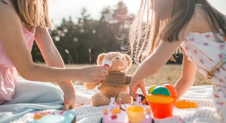 Two girls play with their teddy bears and have a picnic on summer day in park.