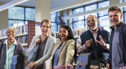A multi-ethnic group of five adults standing in a library, looking up at the camera and smiling.
