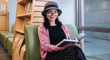 A woman sits reading a book at Dean Drysdale Library. A bookshelf with Grab & Go bags can be seen behind her.