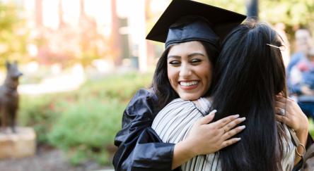 A female graduate in black cap and gown hugs her mother on graduation day.