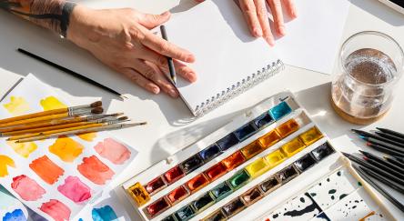Artist starting to draw in sketchbook with watercolour paints off to the side of the image.