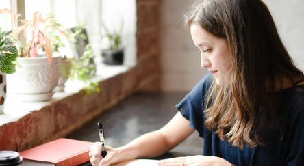 Woman writing in a notebook at a desk by the window.