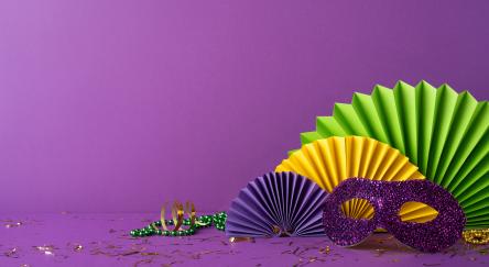 Side view colorful Mardi Gras accessories including a glittery purple mask, festive beads, and paper rosettes arranged on a bold purple background, capturing the celebratory essence of the festival.