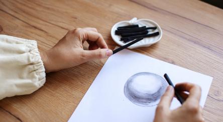 A close-up view of an artist's hands as they skilfully draw a shaded sphere on a white paper using charcoal.