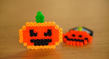 A close-up of two pumpkin shaped Perler charms lying side-by-side on a flat surface.