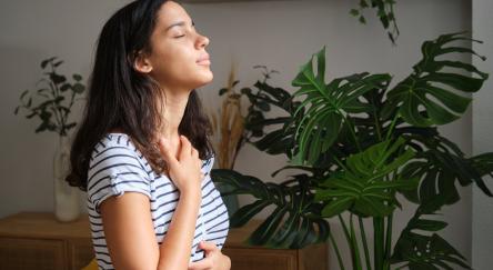 Young woman is taking a deep breath while standing in her living room, surrounded by plants.