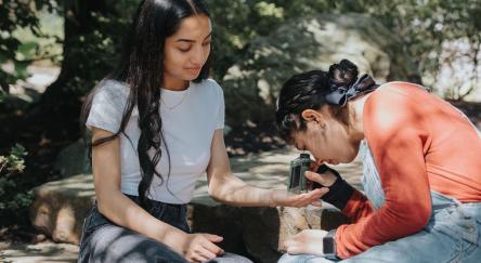 Two teen girls look at bugs and leaves outside through a pocket microscope.