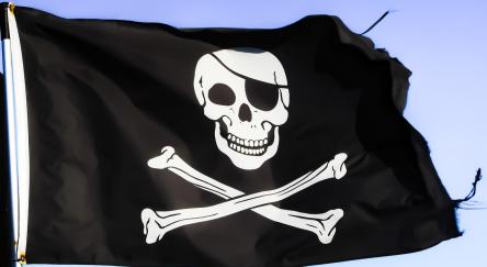 Black pirate flag has a white skull and crossbones with a black eyepatch and is rippling against a blue sky. The edges of the flag are slightly ripped.