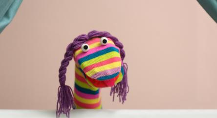 Rainbow sock puppet with purple braids and googly eyes performs against a peach background.