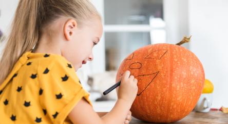 Girl sits at a table drawing a face on a large Halloween pumpkin.