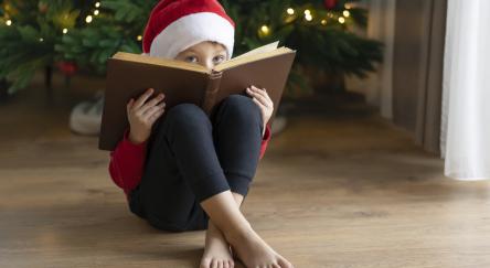 Child in Santa hat peeking over a book. A Christmas tree sits in the background.