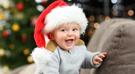 Baby with Santa hat sitting on couch smiling with Christmas tree in the background.