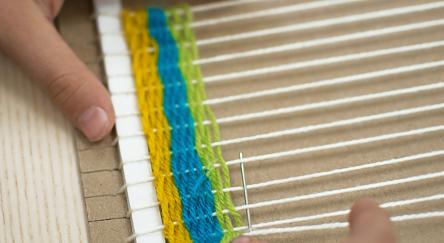 Pair of hands weave colourful yarn through the strings on a cardboard loom.