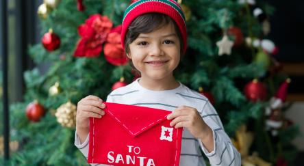 Boy holding a letter for Santa next to the Christmas Tree and looking at the camera smiling.