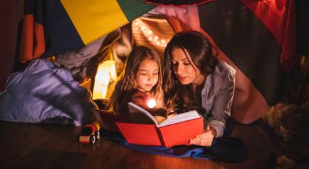 Mother reading a fairy tale book to her daughter under children's blanket fort at night.