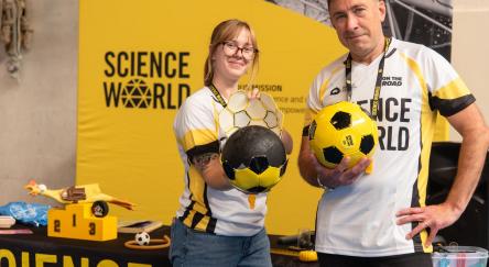 A man and woman holding soccer balls. They are standing in front of a Science World banner.