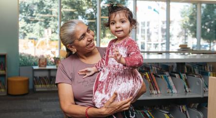 A South Asian woman poses for a photo. She holds her granddaughter, a toddler, in her arms. They have just arrived for Storytime at Terry Fox Library.