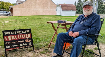 Bearded man sits in a chair with a table. Sign beside him says "I will listen."
