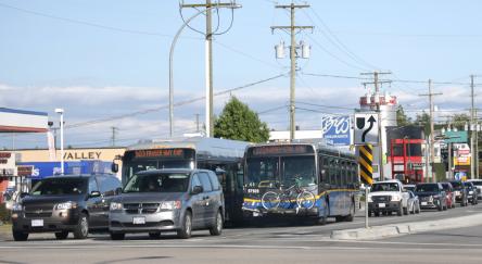 Westbound traffic waits at the intersection at the Fraser Highway and Langley Bypass on a summer afternoon. The TransLink 503 Fraser Hwy Exp and 320 Surrey Ctrl Stn buses travel to Surrey Central Station. One bus carries bicycles.