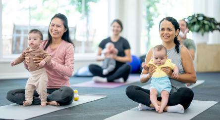 A small group of new parents sit on yoga mats in a studio, with their babies on their laps, as they participate in a parent & baby fitness class.