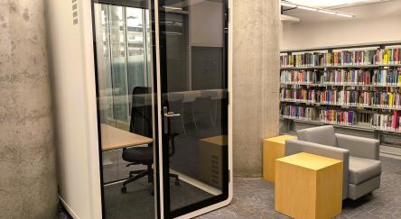 A small, glass-walled privacy booth with a chair and desk inside a modern library space.