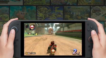 Hands holding a gaming console displaying a racing game on the screen. A character is driving on a dirt track, positioned in second place.