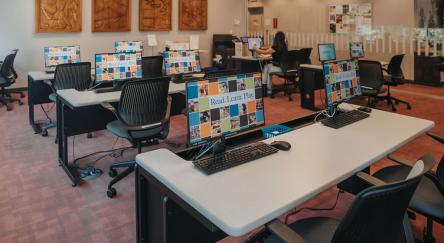 A library computer lab with rows of gray desks, black office chairs, and desktop computers. The screens display a colorful mosaic background with the text "Read. Learn. Play."