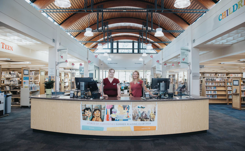 Two women at the information desk of Clearbook Library engage with patrons in a friendly library setting.