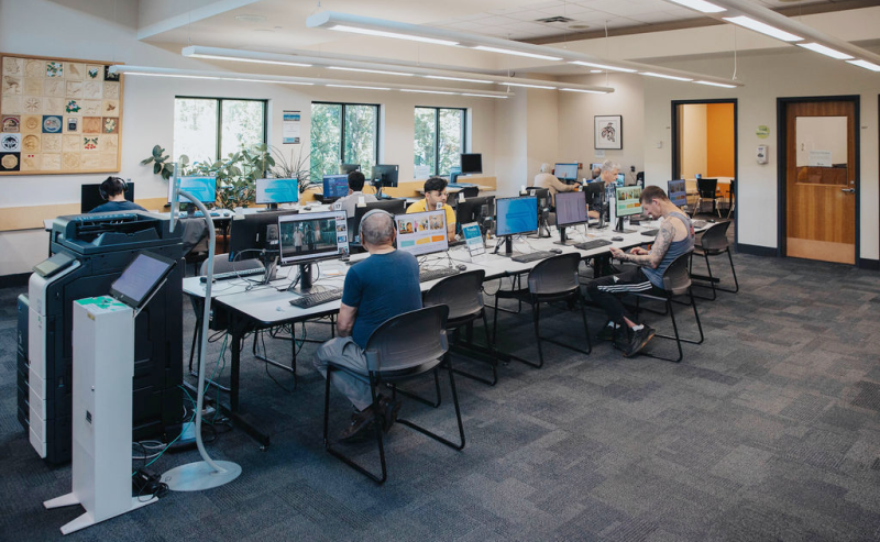 A busy library room with people using computers, large windows letting in natural light.