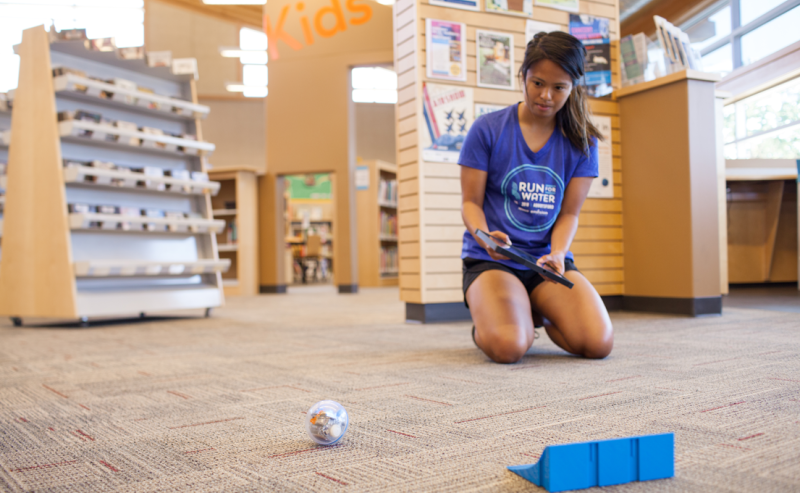 A woman wearing a blue shirt plays with a robot in the Abbotsford Community Library's interior space.