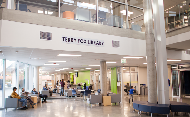 The first-floor lobby of Terry Fox Library, filled with people at tables and others strolling through the lively space.