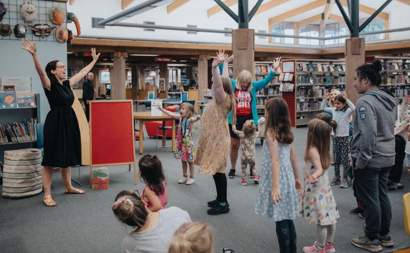 Storytime in the library with a large group reaching about their heads and stretching.