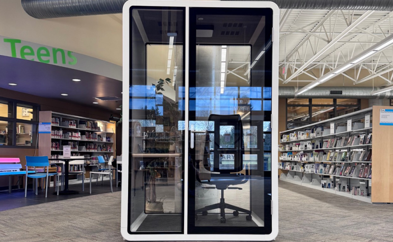 A small, glass-walled privacy booth with a chair and desk inside a modern library space.