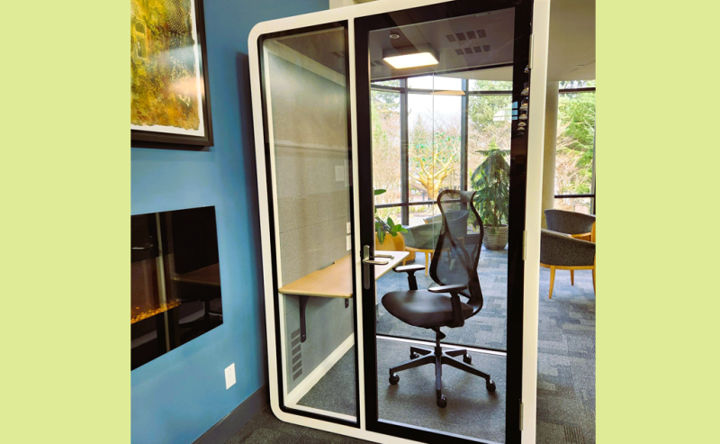 A small, glass-walled privacy booth with a chair and desk inside a modern library space.