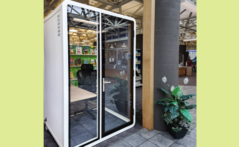 A small, glass-walled privacy booth with a chair and desk inside a modern library space.