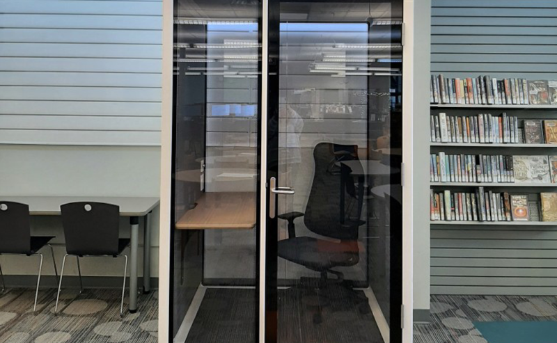 A small, glass-walled privacy booth with a chair and desk inside a modern library space.