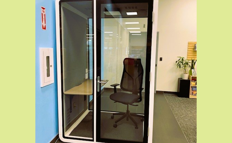 A small, glass-walled privacy booth with a chair and desk inside a modern library space.