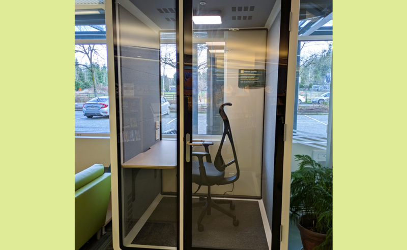 A small, glass-walled privacy booth with a chair and desk inside a modern library space.