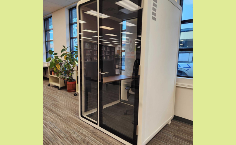 A small, glass-walled privacy booth with a chair and desk inside a modern library space.
