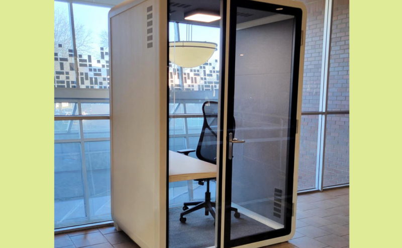 A small, glass-walled privacy booth with a chair and desk inside a modern library space.