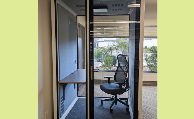 A small, glass-walled privacy booth with a chair and desk inside a modern library space.