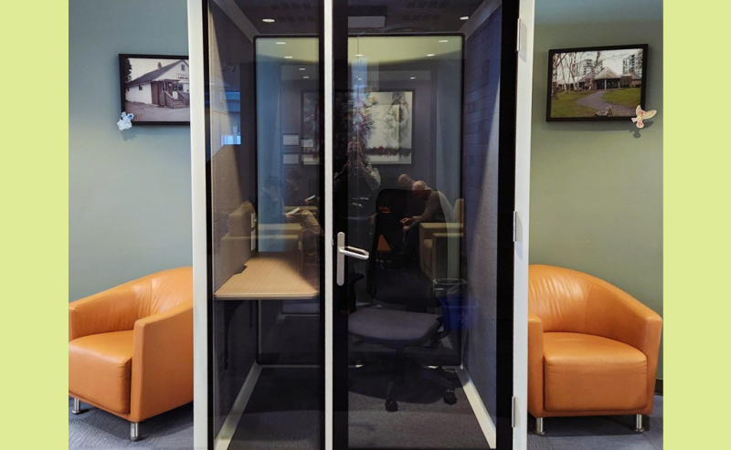 A small, glass-walled privacy booth with a chair and desk inside a modern library space.