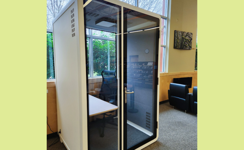 A small, glass-walled privacy booth with a chair and desk inside a modern library space.